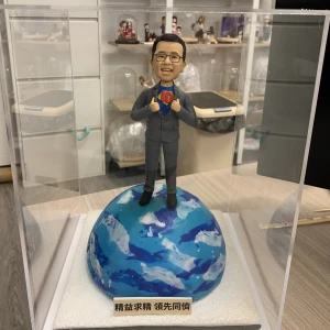 透明展示盒內的人像公仔:短髮戴眼鏡的男士穿深灰西裝,雙手做掀開西裝的動作,露出內層藍色上衣與紅黃色「10」字樣徽章,象徵十周年慶。公仔站在藍白相間的地球模型上,底座前方貼有金色標牌,刻有「精益求精 領先同儕」字樣。背景為工作室環境與層架擺設。整體呈現專業且富儀式感的公司周年禮物擺設。
