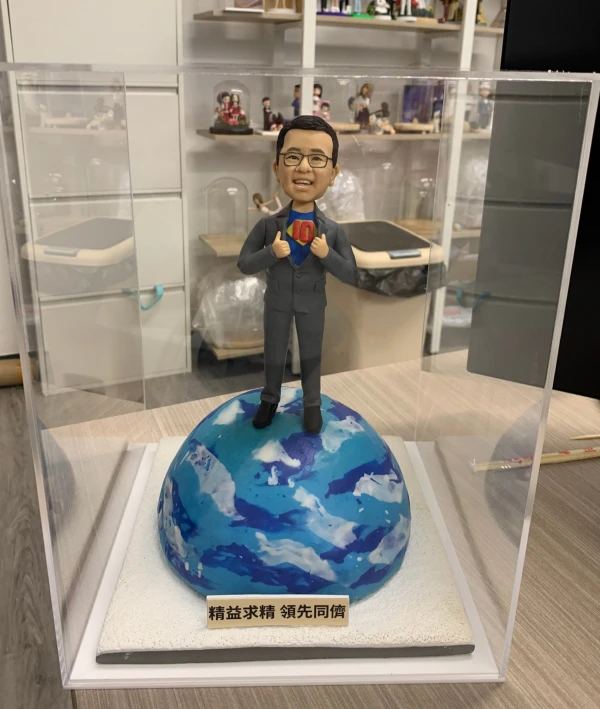 透明展示盒內的人像公仔：短髮戴眼鏡的男士穿深灰西裝，雙手做掀開西裝的動作，露出內層藍色上衣與紅黃色「10」字樣徽章，象徵十周年慶。公仔站在藍白相間的地球模型上，底座前方貼有金色標牌，刻有「精益求精 領先同儕」字樣。背景為工作室環境與層架擺設。整體呈現專業且富儀式感的公司周年禮物擺設。