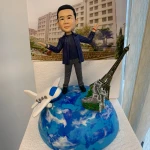 旅遊主題人像公仔生日擺設：短髮男子公仔穿深藍條紋外套、深色上衣與灰褲，張開雙臂站在藍白相間的地球模型上；地球左側有白色藍窗的飛機模型，右側佈置巴黎鐵塔與萬里長城縮景。背後立有建築物照片作為背景板，底座前方貼有祝福字牌。整體呈現以世界旅行慶祝同事生日的人像公仔作品。