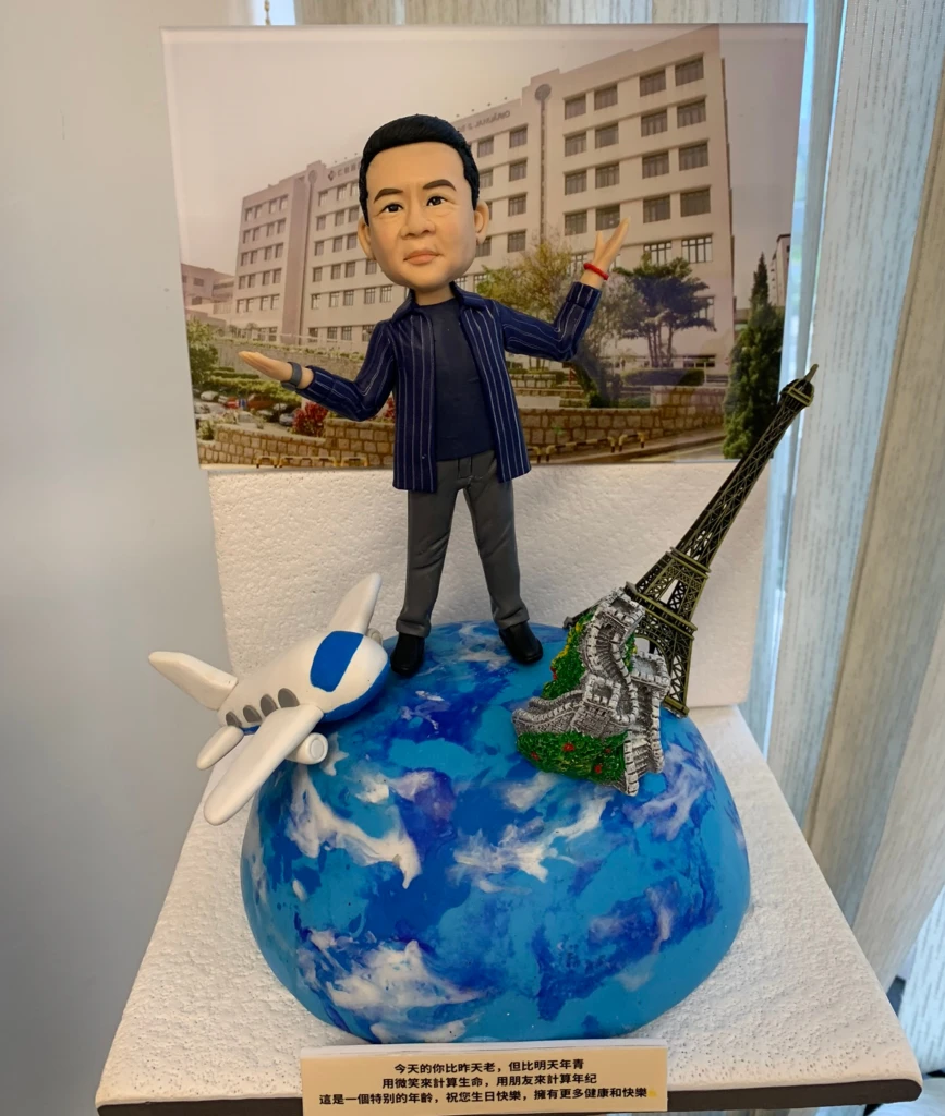 旅遊主題人像公仔生日擺設：短髮男子公仔穿深藍條紋外套、深色上衣與灰褲，張開雙臂站在藍白相間的地球模型上；地球左側有白色藍窗的飛機模型，右側佈置巴黎鐵塔與萬里長城縮景。背後立有建築物照片作為背景板，底座前方貼有祝福字牌。整體呈現以世界旅行慶祝同事生日的人像公仔作品。