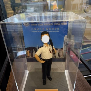 這是一款精心定製的香港警察專屬人像公仔，身穿輔警制服，肩上配有步槍，細節處理極為逼真，從警徽到制服線條均一絲不苟。公仔被安置於透明展示盒中，背景是印有「香港輔助警察隊」字樣與香港天際線的設計，突顯警隊守護社區的責任與使命。整體造型既專業莊嚴，又帶有收藏與紀念的意義，成為一份別具心意的專屬禮物。