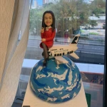 空姐主題的人像公仔坐在迷你飛機上，飛機停靠於藍白相間的地球造型球體上；公仔穿紅色制服外套與裙裝、黑絲襪與高跟鞋，頸繫絲巾、胸前有胸章，右手比出V字手勢微笑示意。飛機機身可見「CATHAY PACIFIC」字樣與綠白尾翼標誌。底座為白色方形平台，背景是窗外城市景與樹木。整體具有旅航氛圍，適合空姐退休的訂製人像公仔退休禮物。