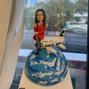 空姐主題的人像公仔坐在迷你飛機上，飛機停靠於藍白相間的地球造型球體上；公仔穿紅色制服外套與裙裝、黑絲襪與高跟鞋，頸繫絲巾、胸前有胸章，右手比出V字手勢微笑示意。飛機機身可見「CATHAY PACIFIC」字樣與綠白尾翼標誌。底座為白色方形平台，背景是窗外城市景與樹木。整體具有旅航氛圍，適合空姐退休的訂製人像公仔退休禮物。