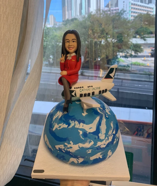 空姐主題的人像公仔坐在迷你飛機上，飛機停靠於藍白相間的地球造型球體上；公仔穿紅色制服外套與裙裝、黑絲襪與高跟鞋，頸繫絲巾、胸前有胸章，右手比出V字手勢微笑示意。飛機機身可見「CATHAY PACIFIC」字樣與綠白尾翼標誌。底座為白色方形平台，背景是窗外城市景與樹木。整體具有旅航氛圍，適合空姐退休的訂製人像公仔退休禮物。