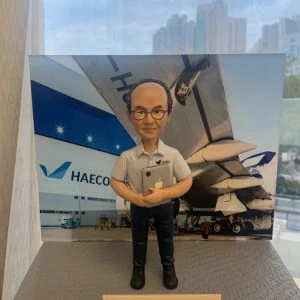 人像公仔退休禮物：中年男士公仔留短髮、戴黑框眼鏡，穿淺灰色短袖襯衫（左胸口繡有 HAECO 標誌）與深色長褲、黑色工作靴，雙手抱著貼有蘋果貼紙的平板電腦，站在灰色質感底座上；前方金色名牌印有「Congratulations on your 50 years of service / So Yiu Bun」。背板為停機坪與大型客機機翼、機庫外牆與 HAECO 字樣的場景，展現專業維修或營運背景。作品於室內靠窗拍攝，畫面下方可見手掌托住底座。