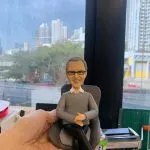 CEO主題人像公仔：灰白短髮、戴眼鏡的男士公仔坐在深灰色紋理單人椅上，雙手自然交疊放於膝上、雙腿交叉，穿淺灰色針織上衣與黑色長褲、黑色皮鞋，面帶微笑。畫面於室內靠窗拍攝，窗外可見高樓大廈與街景；前景有手掌托住座椅，桌面上可見文具與工具罐，呈現寫實辦公氛圍，適合作為老闆或CEO的退休禮物人像公仔。