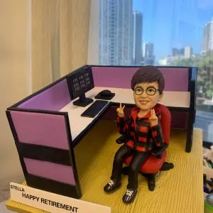 辦公室人像公仔榮休禮：短髮、配圓框眼鏡的上層員工公仔坐在紅色辦公椅上，微笑並以雙手比出雙V手勢；穿紅黑格紋連帽外套、黑色長褲與運動鞋。個人工位為L形配置，粉紫色隔板、白色桌面，配有雙螢幕（畫面顯示「FTE」字樣）、鍵盤、滑鼠與黑色桌上型電話；地台為淺木紋質感。前方白色名牌印有「STELLA｜HAPPY RETIREMENT｜YOUR GS COLLEAGUES｜2023」。作品於室內靠窗拍攝，窗外是晴朗藍天與城市高樓，整體呈現活力、親切且富儀式感的榮休主題辦公室人像公仔。