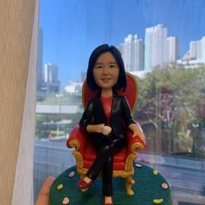 辦公室主題的人像公仔退休禮物：短髮女老闆公仔面帶微笑，穿黑白花紋無袖上衣，坐在黑色辦公椅後、深棕色木質辦公桌前。桌面配置黑色鍵盤、滑鼠、雙螢幕與桌燈，桌側有白色複合式影印機/印表機細節。背景為辦公室玻璃隔間與百葉窗，地台為灰色質感地面，前方小名牌印有「Morgan Stanley」。整體封裝成場景式模型，於室內靠窗拍攝，窗外可見高樓城市景。作品呈現專業而溫暖的榮退氛圍，適合作為老闆的退休禮物人像公仔。