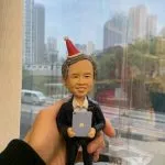 人像公仔退休禮物：中年至長者男士公仔留短灰髮、面帶微笑，頭戴紅白相間的派對尖帽；身穿深色西裝外套與白襯衫，雙手抱著帶有蘋果圖案的小平板電腦。公仔站在灰色石板紋底座上，由手掌托起於室內靠窗拍攝，窗外可見城市高樓與道路景致；畫面下方有辦公室文具與展示物，呈現輕鬆又正式的慶祝氛圍，適合作為老闆的退休禮物人像公仔。