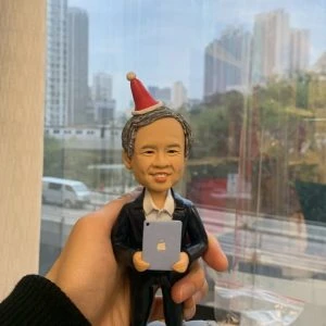 人像公仔退休禮物：中年至長者男士公仔留短灰髮、面帶微笑，頭戴紅白相間的派對尖帽；身穿深色西裝外套與白襯衫，雙手抱著帶有蘋果圖案的小平板電腦。公仔站在灰色石板紋底座上，由手掌托起於室內靠窗拍攝，窗外可見城市高樓與道路景致；畫面下方有辦公室文具與展示物，呈現輕鬆又正式的慶祝氛圍，適合作為老闆的退休禮物人像公仔。