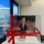 企業主題的人像公仔退休禮物：短髮男士公仔身穿深色西裝外套、白襯衫與紅色領帶，右手拿著一瓶紅色標誌的小汽水；公仔站在圓形白色底座上，底座前方名牌刻有「Keith Fung｜Happy Retirement 2023」。背景板為紅白相間的品牌展示牆與辦公室休憩區場景，前景以透明壓克力展示盒包裹，外繫紅色緞帶與金字「CUTEFIGURE HK」綁帶蝴蝶結；作品由手掌托起拍攝，畫面左側可見大面窗景與城市建築。