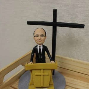 一款客製化的牧師人像公仔，呈現牧師站在講台後方準備講道，背後有大型十字架，是一份送給牧者的感謝禮物。