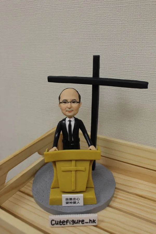 一款客製化的牧師人像公仔，呈現牧師站在講台後方準備講道，背後有大型十字架，是一份送給牧者的感謝禮物。