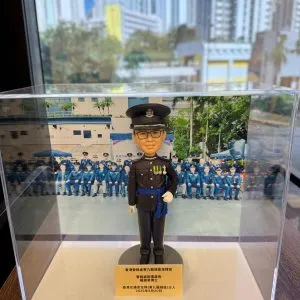 透明壓克力展示盒內擺放一尊以「高級督察」職級為主題的警察人像公仔。公仔頭戴黑色禮帽，帽徽與銀色帽帶清晰；短髮、戴黑框眼鏡，微笑神情。上身著黑色禮服外套，胸前有勳略與綬帶金屬件，肩章與金屬鈕釦立體分明；腰間繫藍色禮儀腰帶，垂掛藍色流蘇。右手自然下垂戴白手套，左手以白手套握於腹前，展現正式儀典姿態；下身為整齊的黑色禮服長褲與黑色禮鞋。公仔立於圓形深灰台座，前方擺放金色名牌，上有致意與日期文字。展示盒內背板是一幅在戶外操場／禮台前的隊列合照，多名著深藍禮服的同袍整齊就座，後方可見校舍或訓練設施與旗桿。展示盒外的環境位於窗邊，窗外是高密度住宅大廈與藍天，室內長條燈具倒影映在玻璃上；整體呈現典禮後的紀念展示與贈禮情境。
