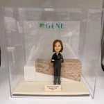 「人像公仔展示於透明盒內，背景為『GENE 皮膚再生專科中心』標誌，公仔身穿黑色西裝、雙手抱胸站立，模擬店鋪主任 Melissa Chan 在新店開張的專業形象，適合作為新店開張禮物。」