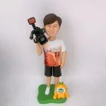 電視台攝錄師主題人像公仔：短髮男子面帶笑容，穿白底橙紅圖案T恤與黑色短褲、白色球鞋，右肩扛專業攝影機，站在綠色草地造型底座上；腳邊有一隻黃色吉祥物小公仔，胸前圓形多色標誌醒目。整體呈現輕鬆又專業的攝錄師形象，適合作為電視台攝錄師退休紀念。