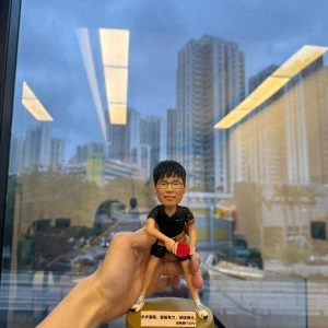 乒乓球比賽主題人像公仔：短髮戴眼鏡的男子公仔身穿黑色運動上衣與短褲，袖口與腰間有金色圖樣點綴；他採低重心準備姿勢，右手握紅色乒乓球拍、左手持白色乒乓球，腳穿白色運動鞋，站在半透明金黃色圓形底座上，底座前貼有中文勵志銘牌。畫面由室內對著落地窗拍攝，可見城市高樓與室內燈光倒影，手持者的手從左側托住公仔。