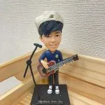 一位反戴畫家帽的男歌手人像公仔，他手持一把低音結他，站在咪高峰架旁，其黑色地台上刻有英文句子 "We Walk With You"。