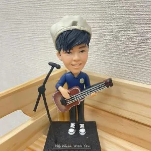 一位反戴畫家帽的男歌手人像公仔，他手持一把低音結他，站在咪高峰架旁，其黑色地台上刻有英文句子 "We Walk With You"。