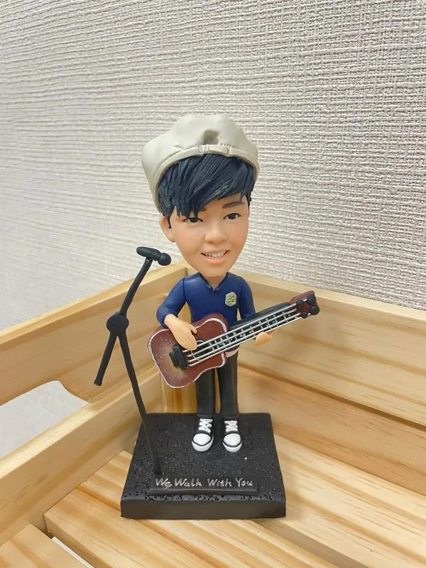 一位反戴畫家帽的男歌手人像公仔，他手持一把低音結他，站在咪高峰架旁，其黑色地台上刻有英文句子 "We Walk With You"。