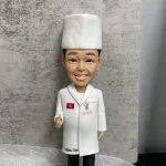 廚師主題人像公仔特寫：戴白色高帽、穿白色廚師袍的男廚師微笑站立，左胸有紅色小旗與三顆星繡章，右胸口有吉祥物圖案；大衣下擺印有「Matsu San」字樣。公仔站在灰色石磚紋底座上，前方白色名牌印「Shikon Yau Yue」。背景為灰色水泥紋牆面與白色紗網質感襯底，前景有綠色底板與黃色、粉色小裝飾。整體呈現隆重且具紀念感，適合作為廚師退休禮物。