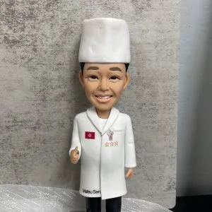 廚師主題人像公仔特寫：戴白色高帽、穿白色廚師袍的男廚師微笑站立，左胸有紅色小旗與三顆星繡章，右胸口有吉祥物圖案；大衣下擺印有「Matsu San」字樣。公仔站在灰色石磚紋底座上，前方白色名牌印「Shikon Yau Yue」。背景為灰色水泥紋牆面與白色紗網質感襯底，前景有綠色底板與黃色、粉色小裝飾。整體呈現隆重且具紀念感，適合作為廚師退休禮物。