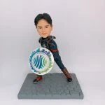 一款英雄主題的COSPLAY人像公仔：短髮少年化身藍黑色戰鬥服造型，胸前有星形標誌與紅色裝飾線，背負肩帶配件，擺出向前衝刺的防守姿勢。左手持圓形盾牌，盾面印有藍綠色地球與城市剪影的圖樣。腳穿黑色鞋與棕色護具，站在灰色六角石紋地台上，背景為簡潔白色。整體呈現活力與守護意象，適合作為COSPLAY 公仔與人像公仔收藏。