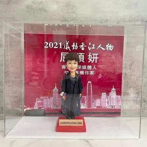 透明亞加力展示盒內的一款精緻手工人像公仔，造型為短髮男子穿深色西裝配黑皮鞋與白恤衫，右手持小花束，站在紅色台座上；台座前方有金色名牌，顯示為香港警隊致贈的禮品（為保護個人識別，名牌人名不展示）。背板為紅色舞台／頒獎設計，上有「2021 感動香江人物」等白色字樣與「資深媒體人、作家」字句，下方繪有香港天際線線條圖。場景置於石紋枱面與灰色牆面前。公仔雕工細膩、五官傳神、塗裝乾淨，整體呈現隆重致敬氛圍。