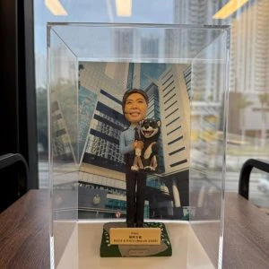 一款祝賀女警務人員晉升的人像公仔禮物，公仔身穿警察制服並懷抱一隻柴犬寵物，背景是警察總部大樓照片。底座銘牌記錄了這次晉升的職級與日期。