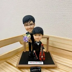 一款音樂老師與學生的雙人訂製人像公仔。小男孩身穿禮服手持大提琴，男老師站在身後拿著花束。公仔地台上有一塊寫上名字的銘牌。