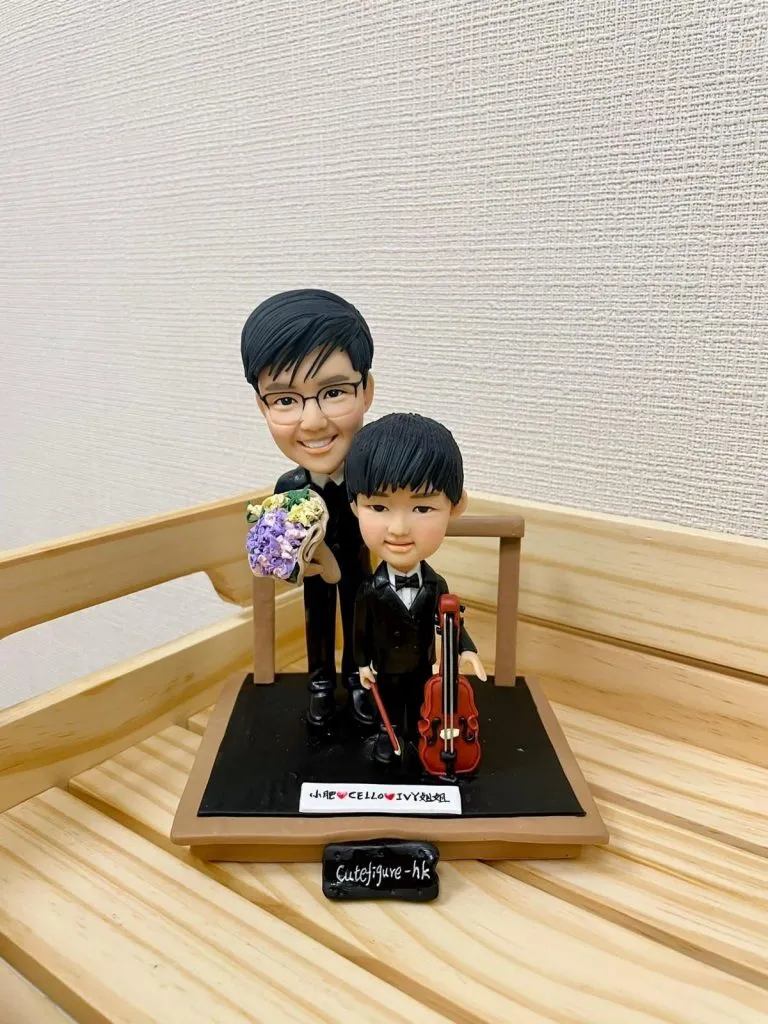 一款音樂老師與學生的雙人訂製人像公仔。小男孩身穿禮服手持大提琴，男老師站在身後拿著花束。公仔地台上有一塊寫上名字的銘牌。