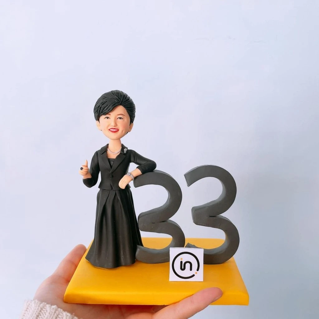 一尊女性商務風人像公仔擺放在亮黃色方形底座上：短髮、微笑，身穿黑色套裝與長裙，胸前別針點綴；右手自然靠在深灰色立體數字「33」上，左手微彎比勢。底座前緣貼有圓角方形小標貼，圖樣似「in」字樣的簡約 Logo。背景為淺色純淨牆面，整體乾淨明亮，呈現適合商務表揚、周年或個人成就紀念的客製人像公仔。