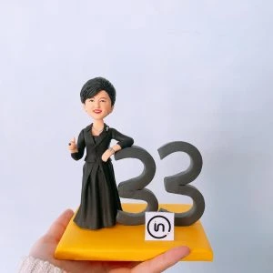 一尊女性商務風人像公仔擺放在亮黃色方形底座上:短髮、微笑,身穿黑色套裝與長裙,胸前別針點綴;右手自然靠在深灰色立體數字「33」上,左手微彎比勢。底座前緣貼有圓角方形小標貼,圖樣似「in」字樣的簡約 Logo。背景為淺色純淨牆面,整體乾淨明亮,呈現適合商務表揚、周年或個人成就紀念的客製人像公仔。