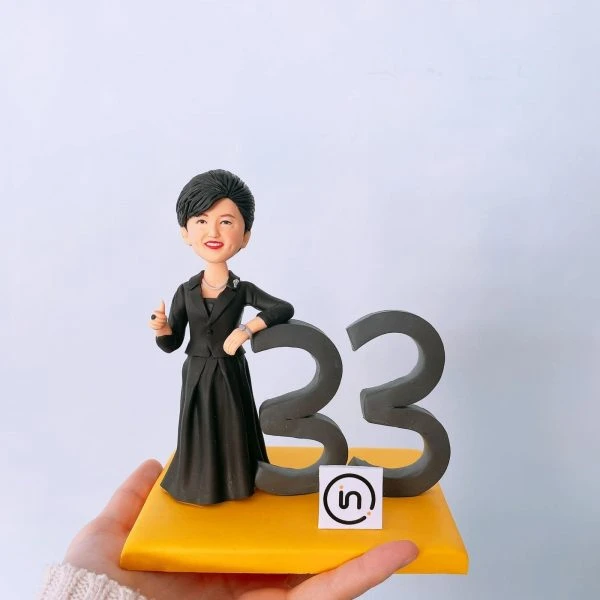 一尊女性商務風人像公仔擺放在亮黃色方形底座上：短髮、微笑，身穿黑色套裝與長裙，胸前別針點綴；右手自然靠在深灰色立體數字「33」上，左手微彎比勢。底座前緣貼有圓角方形小標貼，圖樣似「in」字樣的簡約 Logo。背景為淺色純淨牆面，整體乾淨明亮，呈現適合商務表揚、周年或個人成就紀念的客製人像公仔。