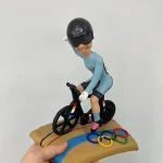 定制單車運動公仔，人物戴黑色安全帽與護目鏡，身穿天藍色緊身運動服，騎著黑色競賽自行車，底座為賽道設計並附有奧運五環彩色裝飾。