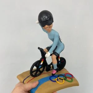 定制單車運動公仔,人物戴黑色安全帽與護目鏡,身穿天藍色緊身運動服,騎著黑色競賽自行車,底座為賽道設計並附有奧運五環彩色裝飾。