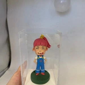 一款客製化的動漫人物人像公仔，一個粉紅頭髮、缺了門牙的淘氣女孩被放置在透明展示盒中，是充滿創意的生日禮物。