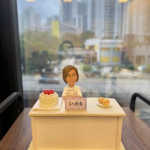 甜品店主題人像公仔擺設：短髮戴眼鏡的女主廚穿白色廚師服，站在淺米色櫃台後方微笑；台面左側放一個覆盆子奶油蛋糕，右側白盤擺放三枚捲蛋糕；正中央立有紫邊小立牌，手寫「玄X廚房」字樣（字跡帶手作感）。整體置於淺黃色底板與木紋桌面上，背後是窗景與高樓大廈的城市背景。造型溫馨且具儀式感，適合作為甜品開張禮物與店內展示。