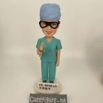 定製醫生人像公仔，角色為外科醫生 Dr. Irene Lo，身穿綠色手術衣與手術帽，配戴護目鏡，手持手術工具，底座寫有「DR. IRENE LO 手術聖手」。此醫護公仔專為外科醫生設計，是獨特的醫療專屬禮物。