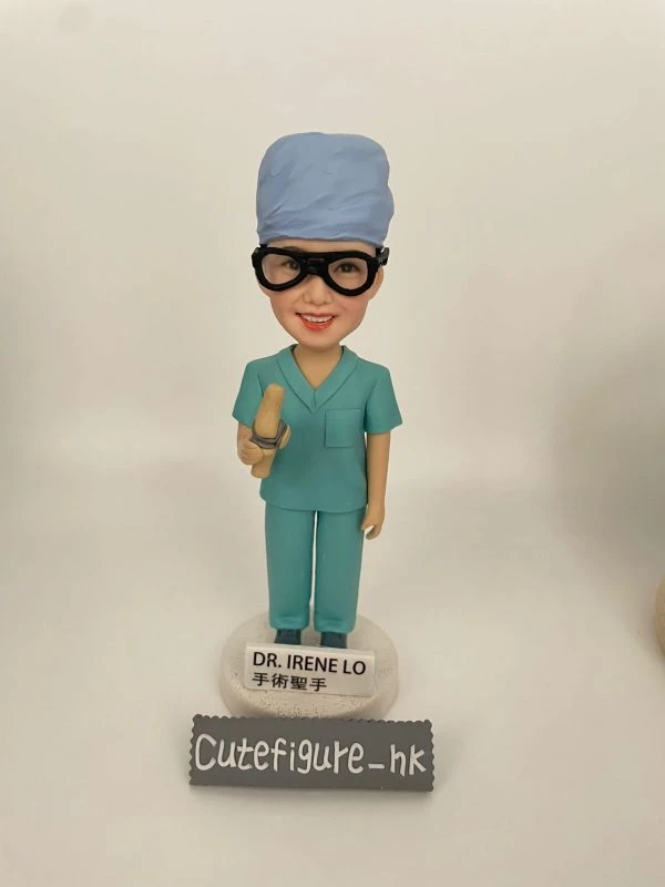 定製醫生人像公仔，角色為外科醫生 Dr. Irene Lo，身穿綠色手術衣與手術帽，配戴護目鏡，手持手術工具，底座寫有「DR. IRENE LO 手術聖手」。此醫護公仔專為外科醫生設計，是獨特的醫療專屬禮物。