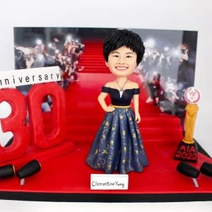 一座慶祝30周年的人像公仔場景：短髮女子微笑站立於紅毯階梯前，穿深藍色露肩禮服，裙擺以金色星月與圖騰點綴，腰間金色腰帶。左側有巨大紅色「30」立體字與「Anniversary」字牌，兩側設黑色欄杆/燈具；右側擺金色獎座，黑紅底座上寫「AIA 2022」。背景是紅毯上媒體攝影群的照片牆，前方白色名牌顯示姓名，整體營造隆重的里程碑慶典氛圍。