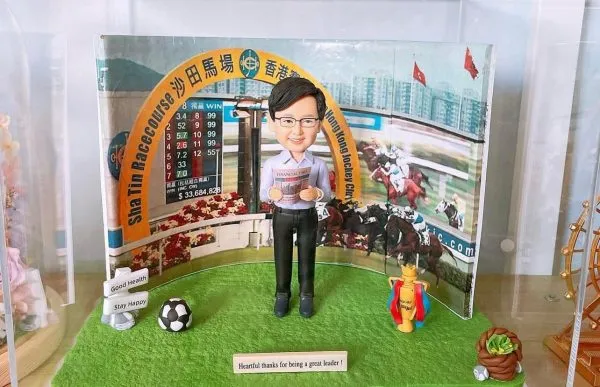 一款為領袖訂製的退休場景人像公仔，公仔手持金融時報，站立在沙田馬場背景前的草地上，底座上擺放著足球、獎盃和祝福語銘牌，以感謝其卓越領導。