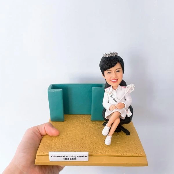 這是一個精緻的手工製作女性護士人像公仔，穿著白色護士制服，頭戴皇冠，手持魔杖。她坐在一張黑色辦公椅上，旁邊有一個綠色的隔板。人像公仔上有一個銘牌，上面寫著“Colorectal Nursing Service, NTEC 2023”。這個公仔展示了一位護士的肖像，應該是代表在結直腸護理領域的醫護專業人士。