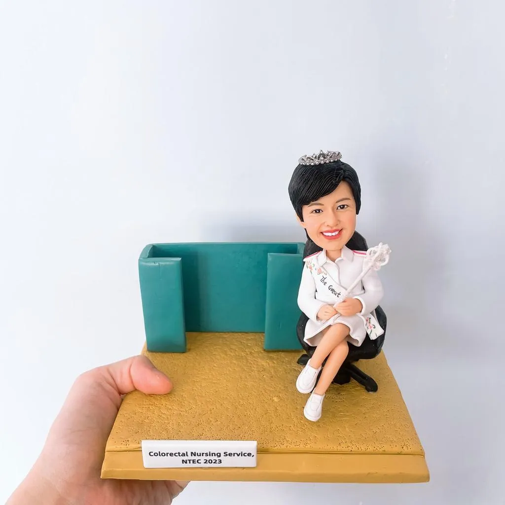 這是一個精緻的手工製作女性護士人像公仔，穿著白色護士制服，頭戴皇冠，手持魔杖。她坐在一張黑色辦公椅上，旁邊有一個綠色的隔板。人像公仔上有一個銘牌，上面寫著“Colorectal Nursing Service, NTEC 2023”。這個公仔展示了一位護士的肖像，應該是代表在結直腸護理領域的醫護專業人士。