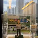 透明壓克力展示盒內，一位男性造型的退休人像公仔站在綠色草地質感底座上，雙手抱胸、面帶微笑，穿淺灰運動外套與深色長褲，腳前踩著一顆黑白相間足球。底座前貼有金色名牌，上刻黑字「感恩有您，同行不懼。」背板是多張足球隊合照拼接而成，穿著不同顏色球衣的學生與教練在球場與球門前合影。展示盒置於窗邊，玻璃外可見高樓大廈天際線的倒影。
