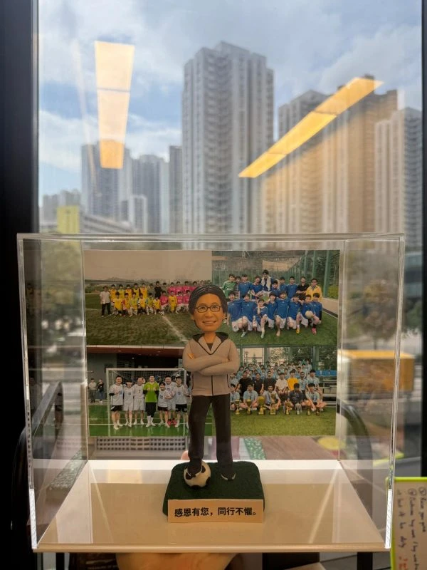 透明壓克力展示盒內，一位男性造型的退休人像公仔站在綠色草地質感底座上，雙手抱胸、面帶微笑，穿淺灰運動外套與深色長褲，腳前踩著一顆黑白相間足球。底座前貼有金色名牌，上刻黑字「感恩有您，同行不懼。」背板是多張足球隊合照拼接而成，穿著不同顏色球衣的學生與教練在球場與球門前合影。展示盒置於窗邊，玻璃外可見高樓大廈天際線的倒影。