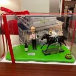 透明展示盒內的退休主題人像公仔場景：戴眼鏡的中年男性公仔身穿粉紅襯衫、黑背心與黑長褲，雙手持香檳與酒杯站立於草地綠底座上，身旁放有一瓶香檳與「Happy Retirement to Albert Lam」英文字樣小牌。右側是一匹黑色賽馬與身穿白色騎師服、黃頭盔的騎師動態奔馳模型，馬匹底座標示「Golden Sixty」字樣；右前角放有綠色蔬果籃小物。背板為現代感建築外牆（Holiday Inn 標誌可見），整體呈現慶祝退休與賽馬主題的收藏級擺設。