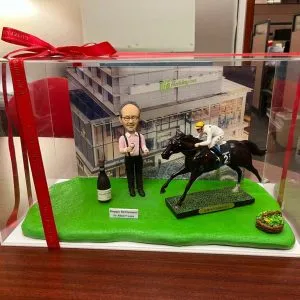 透明展示盒內的退休主題人像公仔場景：戴眼鏡的中年男性公仔身穿粉紅襯衫、黑背心與黑長褲，雙手持香檳與酒杯站立於草地綠底座上，身旁放有一瓶香檳與「Happy Retirement to Albert Lam」英文字樣小牌。右側是一匹黑色賽馬與身穿白色騎師服、黃頭盔的騎師動態奔馳模型，馬匹底座標示「Golden Sixty」字樣；右前角放有綠色蔬果籃小物。背板為現代感建築外牆（Holiday Inn 標誌可見），整體呈現慶祝退休與賽馬主題的收藏級擺設。