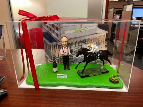 透明展示盒內的退休主題人像公仔場景：戴眼鏡的中年男性公仔身穿粉紅襯衫、黑背心與黑長褲，雙手持香檳與酒杯站立於草地綠底座上，身旁放有一瓶香檳與「Happy Retirement to Albert Lam」英文字樣小牌。右側是一匹黑色賽馬與身穿白色騎師服、黃頭盔的騎師動態奔馳模型，馬匹底座標示「Golden Sixty」字樣；右前角放有綠色蔬果籃小物。背板為現代感建築外牆（Holiday Inn 標誌可見），整體呈現慶祝退休與賽馬主題的收藏級擺設。