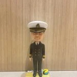 船長主題的人像公仔直立於沙灘質感底座上；戴白色船長帽（黑色帽沿與金色繡飾、帽徽），穿深色雙排扣制服外套與長褲、黑鞋，袖口配金色階級條。底座上點綴貝殼與迷你救生圈等海洋元素。